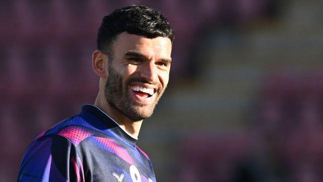 Conor Thomas smiles during a warm-up for Crewe Alexandra. He is wearing a multi-coloured top of black, purple and white.