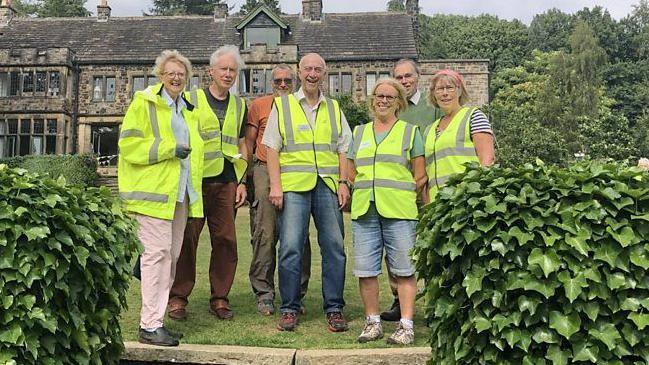 Seven people, stand in a garden with a stone building behind them. They are all smiling and wear yellow high-vis vests.