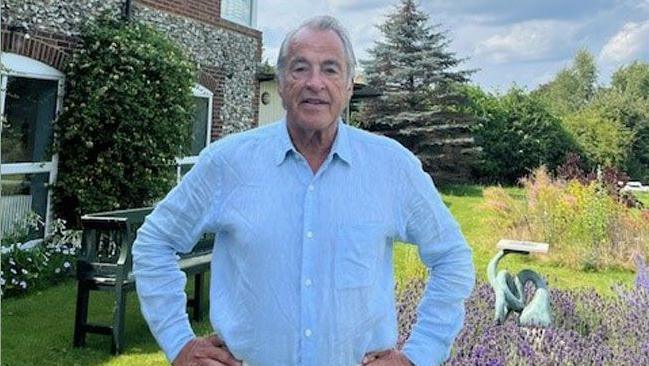 A man wearing a light blue shirt and cream trousers standing with his hands on hips in a garden with a house to his left