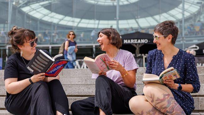 Three women reading books and laughing on the steps of The Forum in Norwich