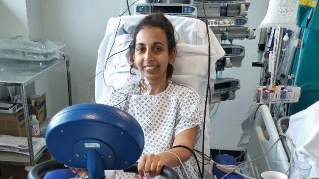 Sanj smiles at the camera, sitting upright in a hospital bed, there are tubes and wires near her face