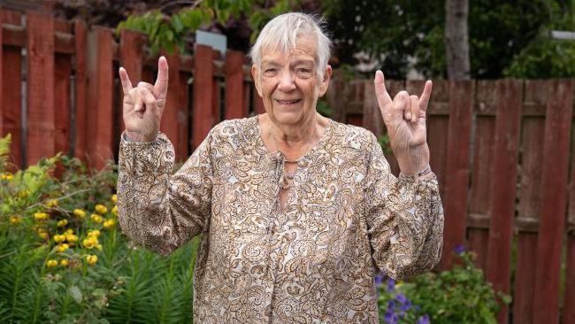 Wilma McKenzie, a woman wearing a top that's brown ad gold, and throwing the devil's horns symbol with both hands.