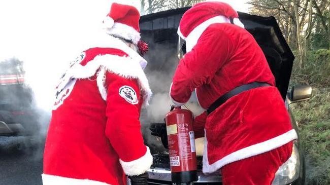 Back view of two people in Santa costumes in front of a car, directing a fire extinguisher into the open bonnet, with clouds of steam or smoke coming from it.