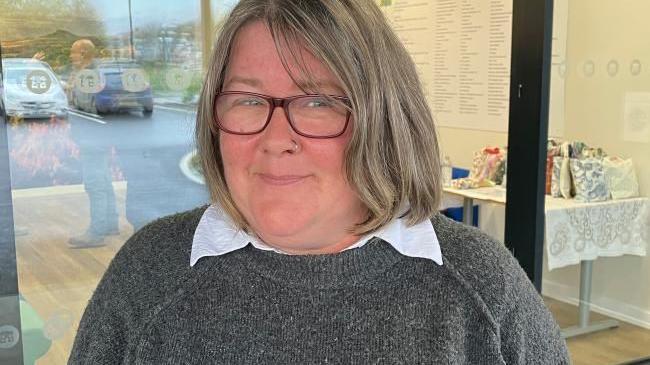 Rhiann stands outside the sliding glass doors of Ryedale Special Families' new centre. She has short grey hair in a bob, black rimmed glasses and a silver nose ring. She wears a grey jumper over a white shirt.