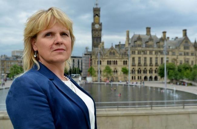 A woman with blonde hair, wearing a white top and dark blue blazer stands slightly to the left of frame with buildings in the background.