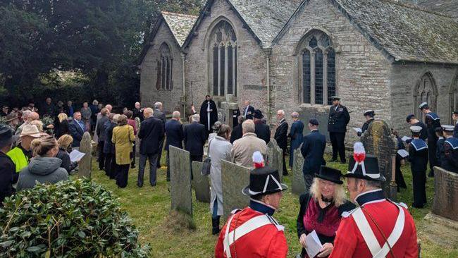 A group of people gathered in a church yard. Some are wearing military dress, others wear coats of differing colours. They are stood amongst grave stones and largely facing away from the camera. There is a church in the background.