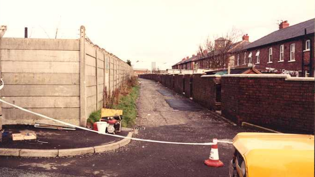 Police cordon across alleyway, between the backs of terraced housing and fences.