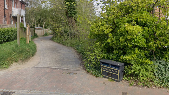 A photo taken from a road looking down a narrow lane with trees on the right. On the left is a brown brick house. There is a large black litter bin on the right of the entrance.