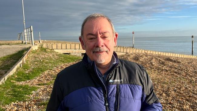 Pedro Cayuela stands on a beach wearing a blue and black coat