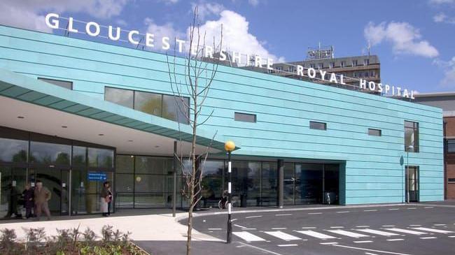 The exterior entrance of Gloucestershire Royal Hospital. It is a large angular building clad in blue horizontal panels. There is a 3D sign on the top of the building and a pedestrian crossing outside the front. It is a sunny day.