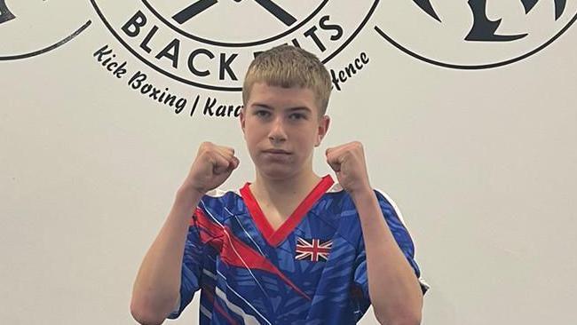 Lewis is wearing a blue, red and white kickboxing uniform. He is holding his fists up at the camera, in a pose. He has short blonde hair.