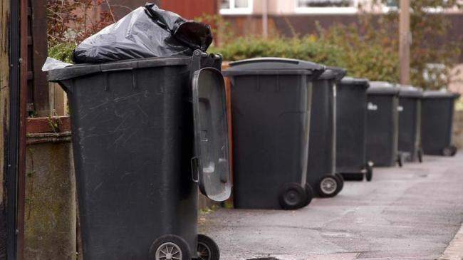 Seven black bins outside houses on a pavement outside properties. The nearest bin is open and a black bin bag is visible at the top.