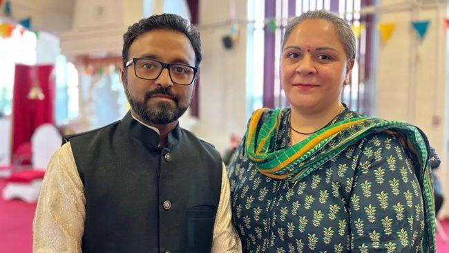 Ashish and Meera standing in a room in the temple, looking directly at the camera. Ashish has short black hair and a neatly trimmed beard and moustache. He has glasses and is wearing a white kurta and a black waistcoat. Meera has tied back grey hair and is wearing a blue and yellow Indian dress with a stole around her neck.