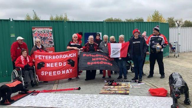 People holding red and black flags with Peterborough Speedway written on them and black Panther sculptures in front of them.