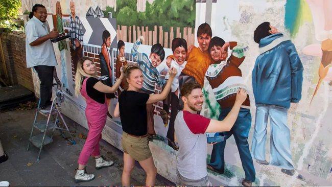 A group of people painting a mural of children playing in a street. They have all turned their heads to smile at the camera. They include a man with black hair standing on a ladder with a paint brush in his hand; a woman with dyed blonde hair wearing pink dungarees and a black shirt; a woman with tied-back blonde hair wearing a black shirt and beige shorts; and a man with brown hair wearing a grey and red top.