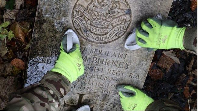 Three hands wearing yellow gloves scrub a gravestone with white brushes. 