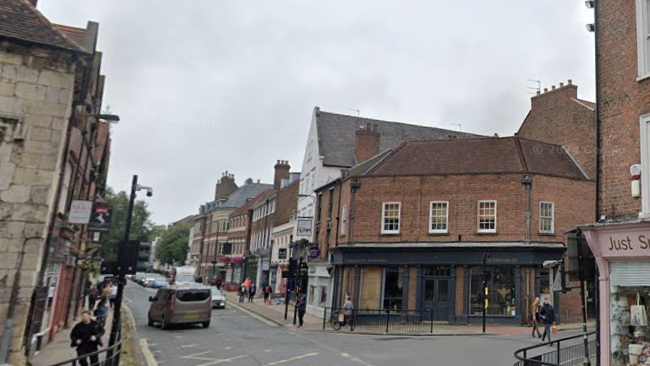 A city centre street junction complete with shop fronts, a yellow box junction, pedestrian crossings and pedestrians walking along the pavements