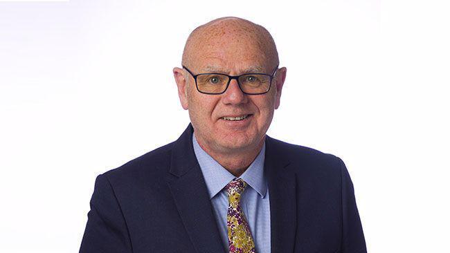 A man in a blue suit jacket and shirt with a floral tie smiles at the camera. He stands in front of a white background.