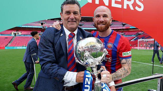 Then Aldershot Town manager Tommy Widdrington in blue suit and red and blue striped tie, with empty Wembley behind, holds FA Trophy