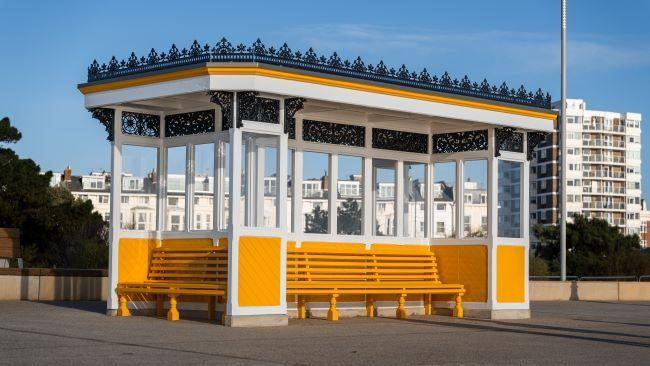 A seafront shelter is pictured on a seafront promenade. It features decorative black ironwork on the roof, with yellow benches and panels underneath. 