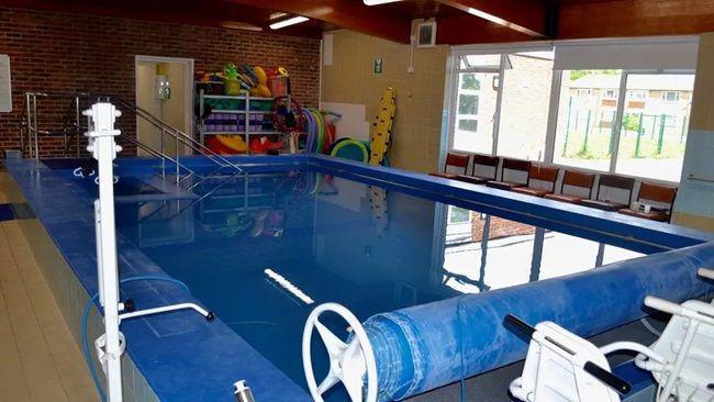 Swimming pool filled with water with cover rolled back. There are chairs and shelves of inflatables next to the pool.