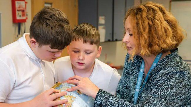 Two schoolboys in white shirts, each with short brown hair, look down at a globe. A woman to the right of the image has her left hand on the globe, with all three looking at where she is pointing. She has wavy blonde hair in a bob hairstyle, and is wearing a turquoise and black patterned shirt. She has a turquoise lanyard around her neck.