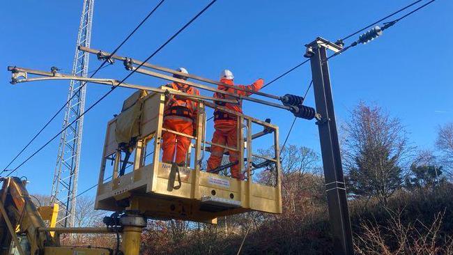 Two engineers in orange overalls and white helmets on a raised platform examining overhead wires