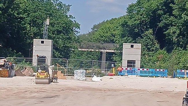 Two rectangular concrete towers on a flat site surrounded by metal railings and blue plastic barriers, with trees behind them