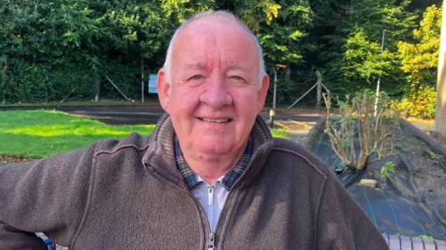 Brian Moore smiling and wearing a brown zip-up cardigan and standing in front of trees