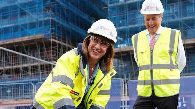 Rachel Reeves has a go at laying bricks on a building site in Birmingham. She smiles at the camera while wearing a hi-vis jacket, gloves, protection googles and a hard hat. The CEO of Berkeley Group, Rob Perrinns, watches on smiling. He is also wearing a hi-vis vest.