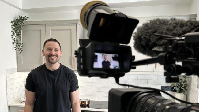 Oliver smiling at a camera, in a kitchen, wearing a black T-shirt, his image is in the camera lens. The kitchen is pale in colour, you can see a microphone with a cover on it, 