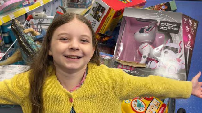 Ivy in a yellow knitted-cardigan with toys in the background. Her arms are outstretched and she is smiling widely. 