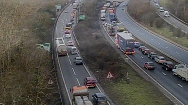 An ariel view from traffic camera of traffic on both sides of the A14, which is dual carriageway. There are wintry trees on the left and bushes on the central reservation.