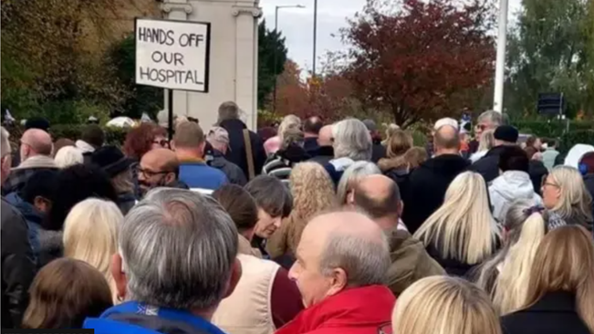 Many people, men and women, gathered outside, surounded by trees. One person holding a placard that reads "hands off our hospital". 