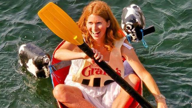 A woman with red hair smiles and sits in a tub in the water and paddles.