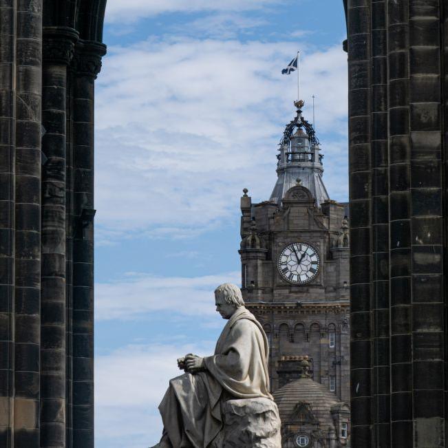 A statue and a clock tower in Edinburgh, framed by a monument, a Saltire flag is flying at the top of the clock tower.
