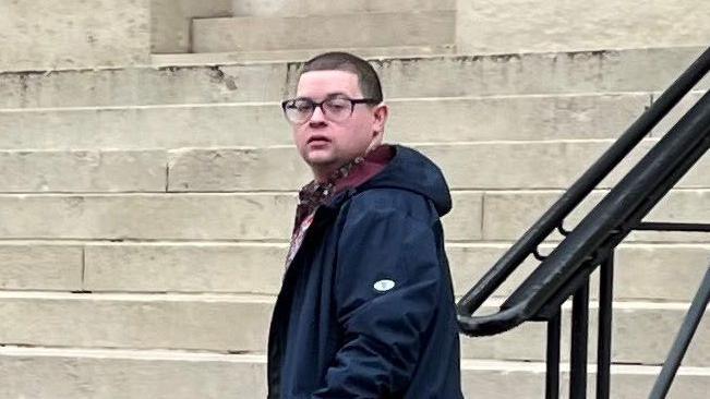 Ieuan Crump wearing a long navy coat and dark glasses, standing in front of steps 