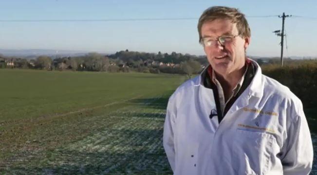 A man standing in front of a field on a farm, wearing a white coat and microphone.
