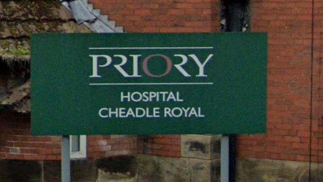 A close up of a green sign set in front of a red brick wall that reads in white writing 'Priory Hospital, Cheadle Royal'