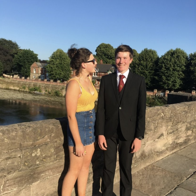 A young woman with her brown hair in a bun stands next to a young man with brown hair. They both smile at each other. She wears a yellow tank top and denim skirt. He wears a black suit and maroon tie. They stand on a bridge over a river.