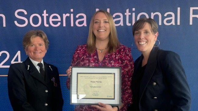 Deputy chief constable Maggie Blyth can be seen on the left wearing her black police uniform with a medal underneath her left lapel. Poppy Murray is positioned in the middle wearing a pink, floral dress while she holds a framed certificate. MP Jess Phillips is standing on the right in a navy blue suit with silver hooped earrings, all are smiling at the camera