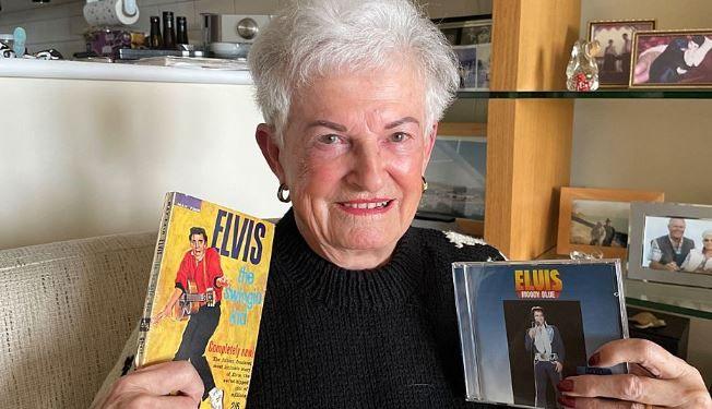 Jan Owen, an older woman, who is sitting indoors, smiling warmly while holding Elvis Presley memorabilia. In her right hand, she holds a book featuring Elvis on the cover with the text "Elvis: The Swingin’ Kid." In her left hand, she holds a CD case titled "Elvis: Moody Blue." The background includes shelves with family photographs and decorations