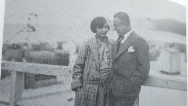 An old black and white photograph of a woman with a dark bob, wearing a long coat, standing talking to a man wearing a suit.