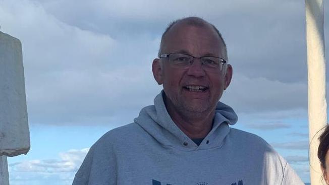 A man in a grey hoodie and glasses with greying hair, smiling at the camera, against a backdrop of blue sky and light grey clouds.