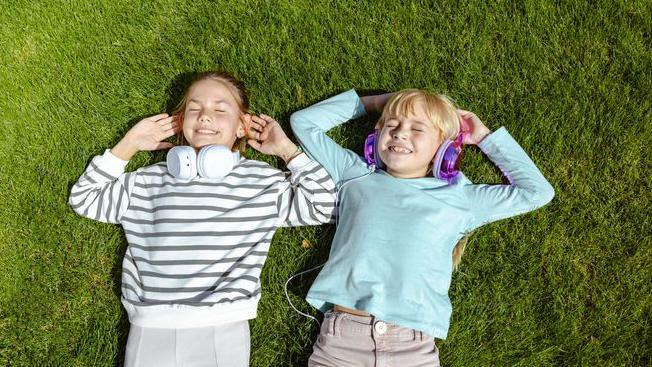 Two young girls lying on grass wearing headphones.