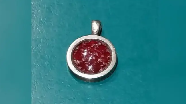 A close-up image of a silver pendant with a red middle. The middle is a dark red in a marble pattern. It is on a teal table. 