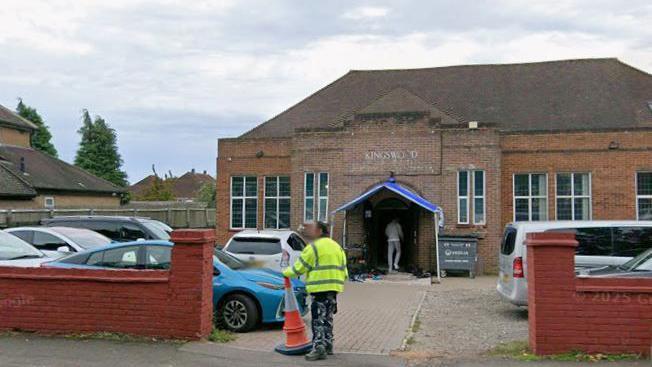 A Google streetview image of the mosque building from outside - with cars parked in front of the building. A person in a yellow high-vis jacket is standing at the entrance to the car park, holding an orange traffic cone.