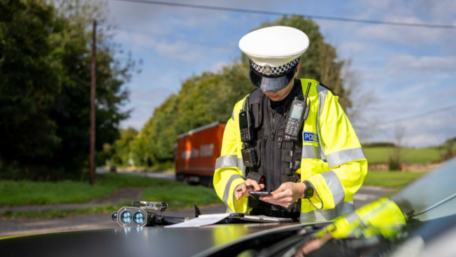 Image shows a police officer looking at their phone and resting again the bonnet of a car. The officer is wearing a police jacket and a bullet-proof vest.