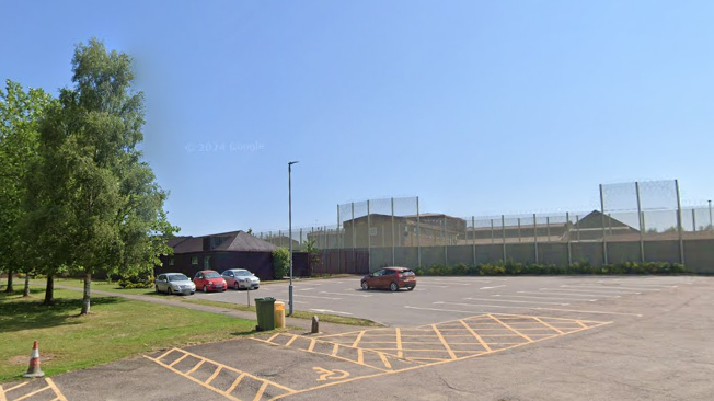 A general view of HMP Huntercombe - which has high metal gates surrounding it - and its car park on a sunny day.