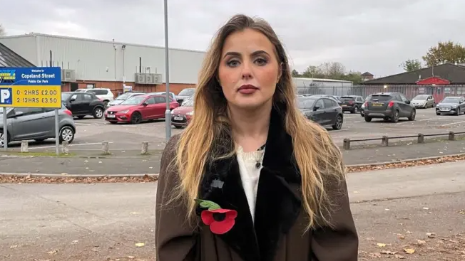 Miss Hudson, who is a young blonde woman, stands outside the Excel carpark in question, wearing a brown coat with a poppy on the lapel